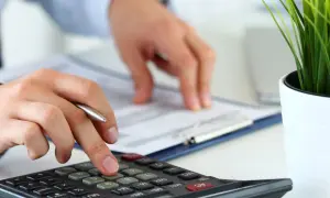 Osoba liczy na kalkulatorze, przygotowując protokół zniszczenia towaru. Obok dokumenty i zielona roślina.