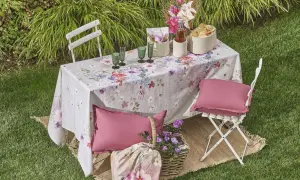 Un tavolo da giardino apparecchiato con una tovaglia floreale, bicchieri verdi, cesti e cuscini rosa. Un'idea per come rivestire un tavolo con stoffa per un picnic.