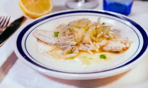 Delicato branzino in padella con cipolle e prezzemolo, servito su un piatto bianco con bordo blu. Accanto, uno spicchio di limone.