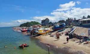 Słoneczny dzień nad polskim morzem. Kolorowe łodzie na plaży, tłumy ludzi, a na wodzie skuter wodny.