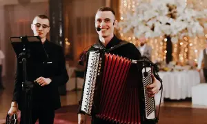 Uśmiechnięty muzyk gra na akordeonie, tworząc radosną muzykę biesiadną. Obok stoi kolega z tamburynem.