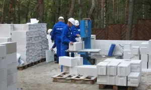 Budowa domu z bloczków betonowych. Pracownicy w niebieskich kombinezonach i białych kaskach układają materiał. Widać piłę do cięcia bloczków.