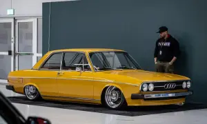 Gul Audi 100 C1, en klassiker med kromade fälgar, står parkerad bredvid en man i keps.