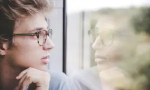 Młody mężczyzna w okularach patrzy w okno, jego odbicie sugeruje głęboką refleksję i inteligencję intrapersonalną.