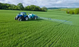 Niebieski traktor z opryskiwaczem na zielonym polu, stosuje środek Chwastox Turbo 340 SL. Dawkowanie jest kluczowe dla skuteczności.