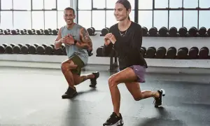 Coppia che esegue affondi, un ottimo esercizio cardio a casa. Sfondo con bilancieri.