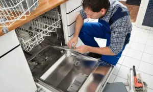 Fachowiec w niebieskim kombinezonie dokonuje podłączenia zmywarki, trzymając w rękach jej element. Obok leżą narzędzia.