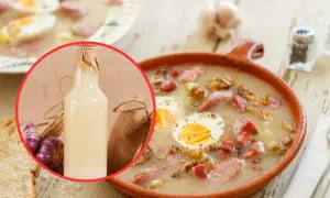 Żurek w misce z jajkiem i kiełbasą, w tle butelka zakwasu, czosnek i chleb.
