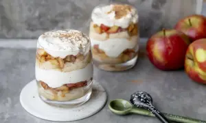 Délicieux tiramisu pomme biscuit cuillère, superposé dans des verres avec de la crème fouettée et de la cannelle. Des pommes rouges sont à côté.