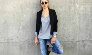 Stylisches Outfit mit Blazer: Frau in Jeans, grauem V-Shirt und Sonnenbrille lehnt an Betonwand.