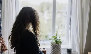 Flicka med långt hår tittar ut genom fönstret. En planta står på fönsterbrädan. Bilden kan symbolisera emotionell utveckling 3 år.