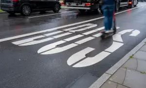 Jazda hulajnogą po buspasie grozi mandatem. Na mokrej drodze widać białe linie wyznaczające pas oraz poruszającego się po nim użytkownika hulajnogi.