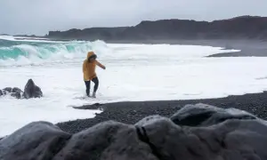 Kobieta w żółtej kurtce brodzi w pianie na czarnej plaży Reynisfjara, podczas gdy potężne fale rozbijają się o brzeg.