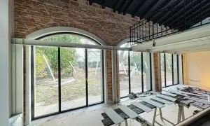 Interno in fase di ristrutturazione con ampie vetrate sul giardino. In primo piano, assi di legno appoggiate su cavalletti, segno di lavori di miglioramento della casa in corso.