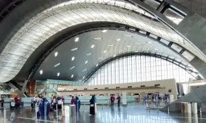 Nowoczesne wnętrze lotniska w Doha z charakterystycznym, falującym dachem i przeszklonymi ścianami. Pasażerowie czekają w kolejce do odprawy.