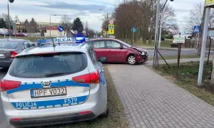 Samochód policyjny z włączonymi sygnałami świetlnymi przy drodze, gdzie doszło do kolizji drogowej. Czerwony samochód ma uszkodzony przód.