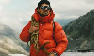 Mężczyzna w jaskrawopomarańczowej kurtce puchowej, czapce i okularach przeciwsłonecznych, z liną i czekana, gotowy na przygodę w stylu gorpcore.