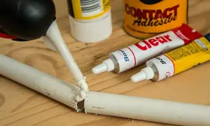 Aplikacja kleju z tubki na pękniętą plastikową rurkę. Obok leżą tubki z klejem i puszka "Contact Adhesive", pokazujące, czym skleić plastik.