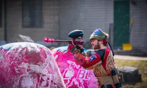 Gracz w stroju bojowym celuje z markerem, gotowy do akcji w laserowym paintballu. Za nim różowe dmuchane przeszkody.
