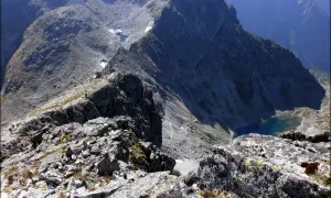 Widok z grani na skaliste zbocza i turkusowe jezioro. Mała, wysoka postać turysty na szlaku.