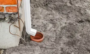 Biała rura spustowa odprowadza wodę do ceramicznej studzienki. Widać fragment ceglanego muru i betonowego cokołu.