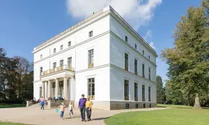 Familie spaziert durch den Jenisch Park vor einem weißen Herrenhaus mit Säulen und goldenen Verzierungen.