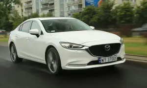 Biała Mazda 6 2.0 benzyna jedzie po mokrej drodze, w tle budynki i drzewa.