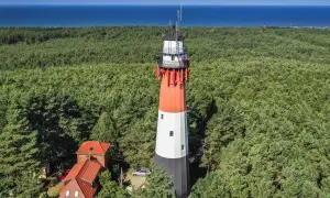 Stylowa latarnia morska w lesie sosnowym, z widokiem na błękitne morze. Obok budynki i samochód.
