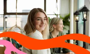 Två leende kvinnor sitter vid ett bord. Den ena tittar mot kameran. Kanske funderar de på vad som går arbetsgivaravgiften till.