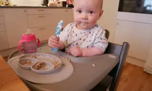 Sött litet barn med mat i ansiktet, redo att introducera mat bebis. En tallrik med mat och en mugg finns bredvid.