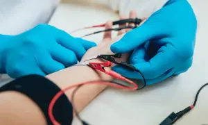 Lekarz w niebieskich rękawiczkach podłącza elektrody do ręki pacjenta. To badanie EMG, które pozwala ocenić funkcję nerwów i mięśni.