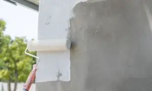 Wałek malarski pokrywa białą farbą szary beton. Prace wykończeniowe na zewnątrz budynku.