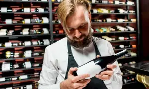 Un sommelier esamina una bottiglia, un passo fondamentale per capire se un vino è buono. Dietro di lui, scaffali pieni di bottiglie.