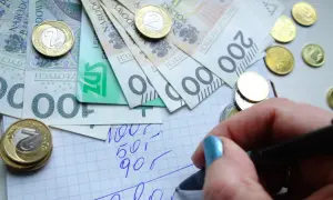 Ręka z niebieskim lakierem pisze na kratkowanym papierze, obok leżą polskie banknoty i monety. Planowanie wydatków to pierwszy krok do rewaloryzacji budżetu.