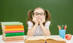 Uśmiechnięta dziewczynka w okularach z kitkami, oparta o ręce, siedzi przy biurku z książkami i jabłkiem. To idealny ipet do nauki.