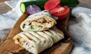 Pyszny kebab w tortilli z kurczakiem, warzywami i sosem, podany na desce z pomidorem i ogórkiem.