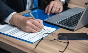 Biegły sądowy w garniturze pisze długopisem na dokumentach. Obok leży laptop, okulary i telefon.