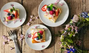 Tre tallrikar med vegansk midsommarmat, toppad med bär och blommor, ligger på ett träbord. En blomsterkrans ligger bredvid.