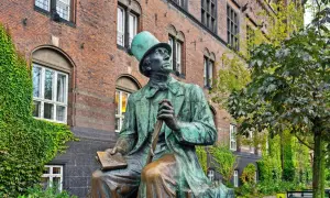 Pomnik Hansa Christiana Andersena w Kopenhadze, siedzącego z książką i laską, na tle ceglanego budynku porośniętego bluszczem.