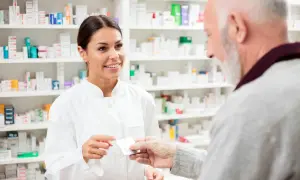 Uśmiechnięta farmaceutka podaje starszemu panu opakowanie leków. W tle apteczne półki pełne medykamentów, tworzące poltixa.