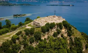 Der Gardasee von oben: Ruinen einer Festung auf einem Hügel mit Kreuz, davor grüne Bäume und im Hintergrund die Inseln und das blaue Wasser.