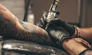 Manos enguantadas trabajan en un tatuaje colorido en un brazo. El artista usa una máquina de tatuar, creando arte permanente.