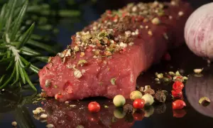 Un pezzo di carne succulenta, condita con pepe misto e rosmarino, pronta per diventare una deliziosa carne tipica pugliese. Accanto, uno spicchio d'aglio.