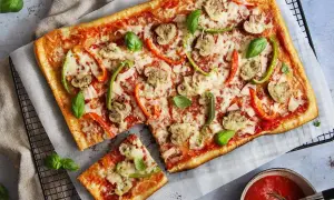 En avskuren bit av en rektangulär pizza med skinka, champinjoner och paprika. Vilken skinka till pizza är bäst? Frågan är svår att svara på, men denna ser god ut!