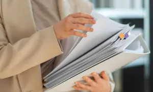 Kobieta w beżowej marynarce przegląda dokumenty. Wśród nich znajduje się wypis z księgi wieczystej, oznaczony kolorowymi zakładkami.