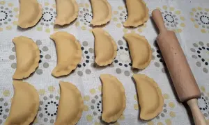 Gotowe do gotowania pierogi z jabłkami, ułożone rzędami na wzorzystym obrusie, obok drewnianego wałka.