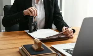 Osoba w garniturze stempluje dokumenty, być może związane z hipoteką. Na biurku leżą też książki i laptop.