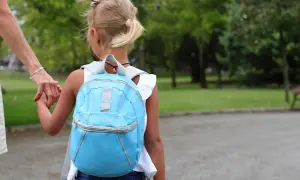 Dziecko w niebieskim plecaku trzyma za rękę dorosłego, idąc przez park.