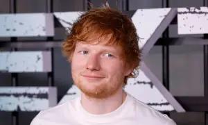 Ed Sheeran w białej koszulce, z rudymi włosami i niebieskimi oczami, uśmiecha się.