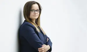 Kobieta w okularach i granatowej marynarce, oparta o białą ścianę.